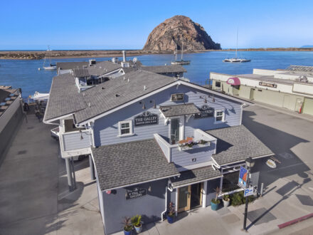 Aerial view of a waterfront area with a gray building housing "The Galley Seafood Grill & Bar" and "Anderson Inn". The building is situated near a calm body of water with boats docked and a large rock formation in the background under a clear blue sky.