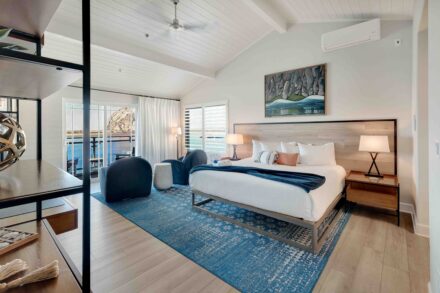 Modern bedroom with vaulted ceiling featuring a bed with white sheets and a wooden headboard. The room has wooden flooring, a blue area rug, and large windows with white curtains allowing ample natural light. Two bedside tables with lamps and two black chairs are placed near the window.