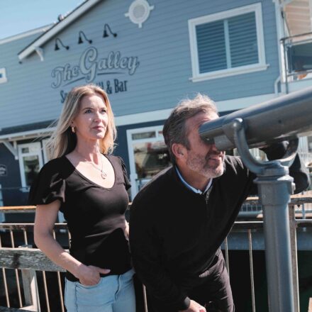 A man looks through a coin-operated binocular viewer while a woman stands beside him looking into the distance. They are on a dock next to a blue-gray building labeled "The Galley Seafood Grill & Bar." The scene appears to be outdoors on a sunny day.