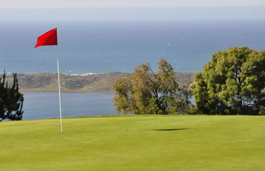 A vibrant red flag marks the hole on a green golf course near the ocean. Behind the course, there's a picturesque view of the blue sea and a small hilly land area. Lush green trees frame the scene on a clear, sunny day.