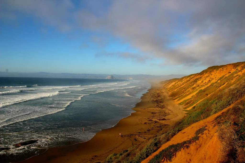 A scenic view of a beach with golden sand dunes on the right, waves gently hitting the shore on the left, and a clear blue sky with scattered clouds above. Vegetation partially covers the dunes, and the horizon features distant land and hills.