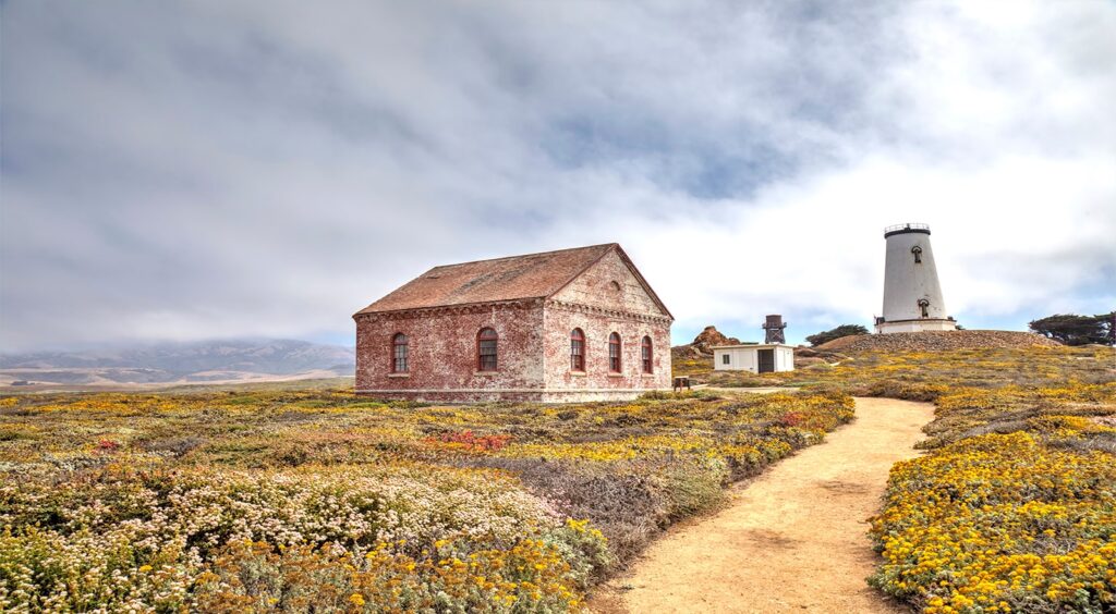 A dirt path leads to an old, brick building and a white lighthouse with a black top, situated on a grassy field dotted with yellow and white wildflowers. In the background, a range of mountains is visible under a partly cloudy sky.