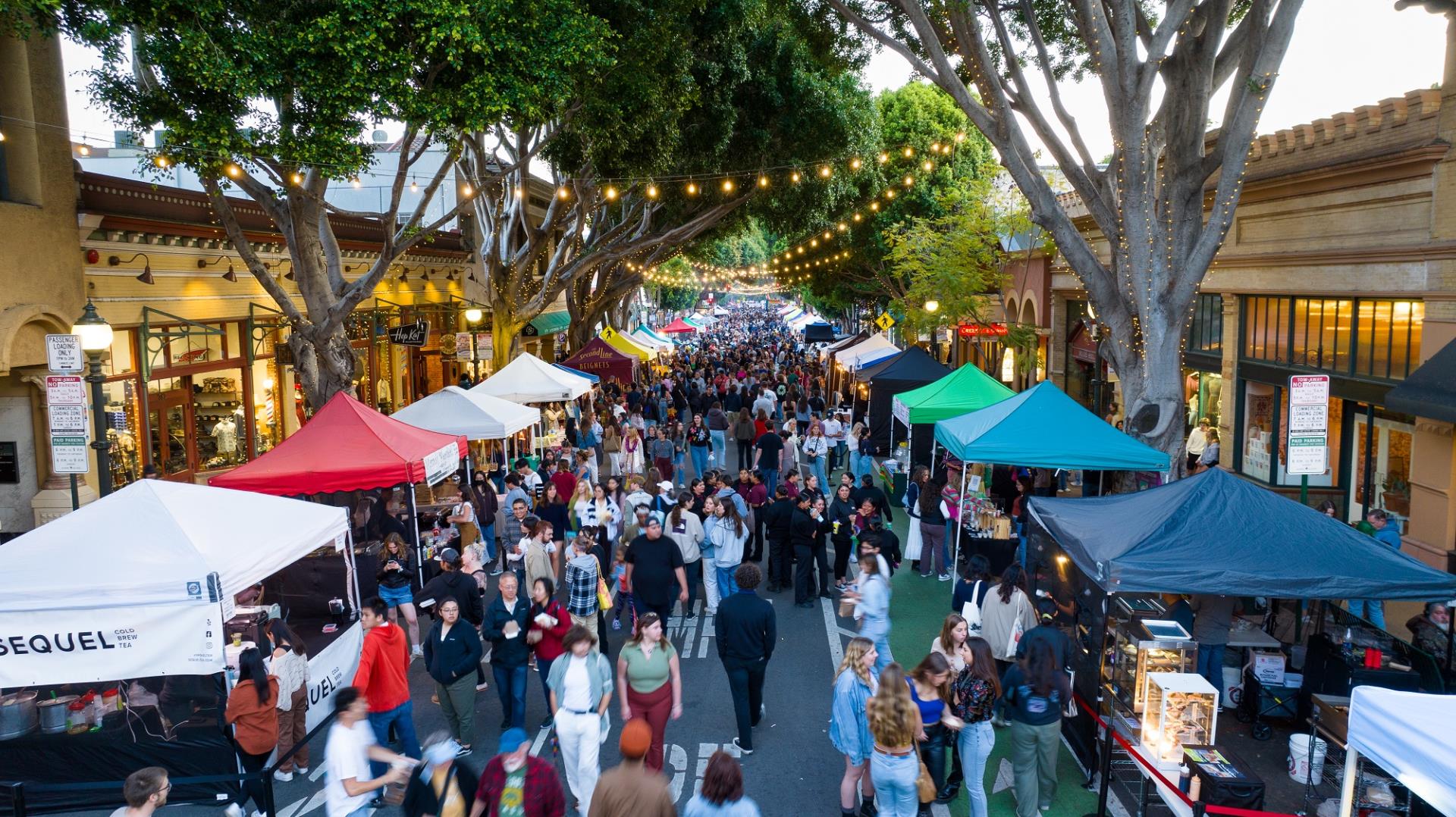 A bustling outdoor street market with crowds of people walking between vendor booths lined along both sides. The booths have colorful canopies, and string lights are hung overhead. Tall trees flank the street, adding a vibrant, festive atmosphere.