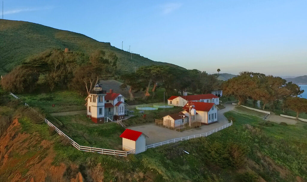 Aerial view of a picturesque coastal scene featuring a quaint lighthouse with an adjacent house, red roofs, and surrounding greenery. The structures are situated on a grassy cliff overlooking the ocean, with a winding path leading through the area.