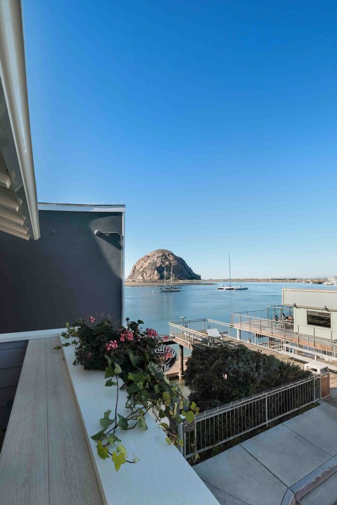 View of a calm bay featuring a large rock formation in the distance. The foreground has a balcony with potted flowers, wooden railings, and buildings on the waterfront under a clear blue sky. Sailboats are anchored in the water.