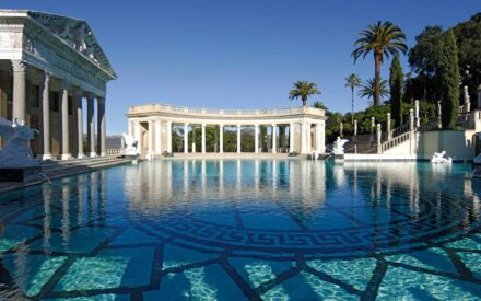 A luxurious outdoor swimming pool with clear blue water, surrounded by marble statues and Greco-Roman style architecture, including columns and ornate structures. Palm trees and lush greenery are visible in the background under a clear blue sky.