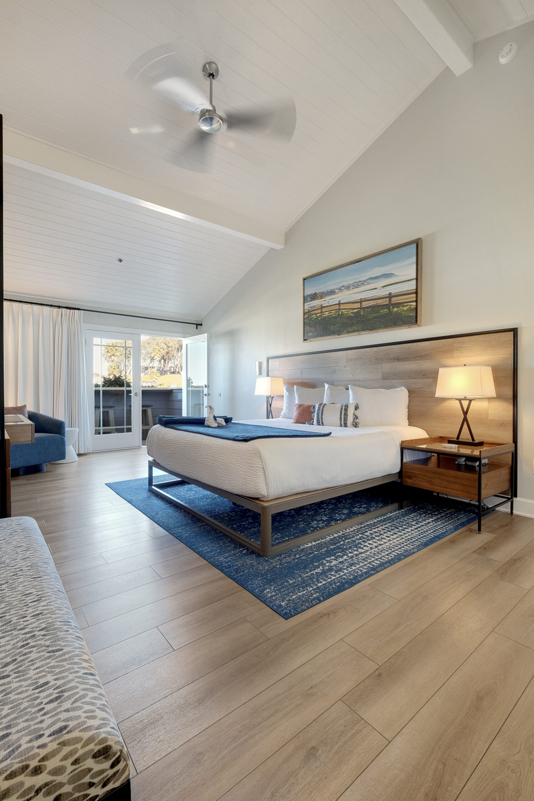 A modern bedroom with a double bed, wood headboard, and blue area rug on light wood floors. The room includes two nightstands with lamps, a ceiling fan, and a large framed landscape print above the bed. A sitting area and a sliding glass door are visible in the background.