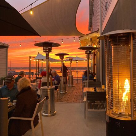 A cozy outdoor restaurant patio at sunset, featuring several patrons dining and sitting near tall standing heaters. The sky is ablaze with orange and pink hues, casting a warm glow over the scene. String lights hang above, enhancing the inviting atmosphere.