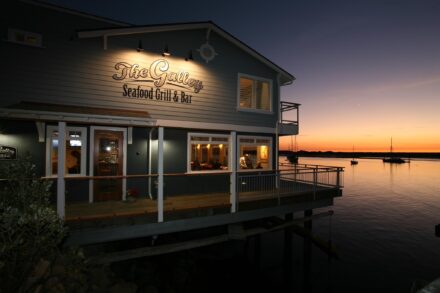 A seaside restaurant named "The Galley Seafood Grill & Bar" over a calm body of water at sunset. The building is illuminated with warm lights, and boats are visible in the background, anchored on the water. The sky displays deep twilight hues.