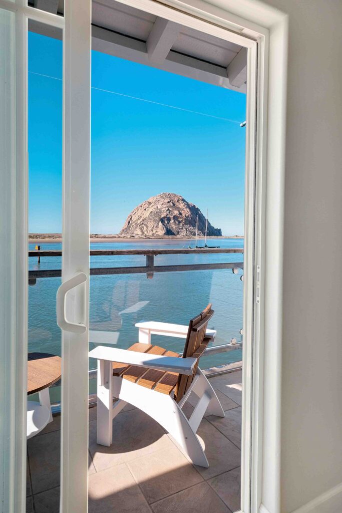 A view through a sliding glass door shows a porch with two wooden Adirondack chairs placed around a small table, overlooking calm waters. In the distance, a large, rounded rock formation rises against a clear blue sky.