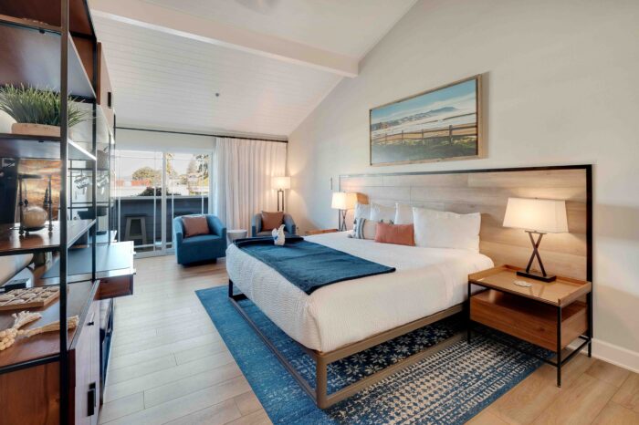 A modern hotel room features a large bed with white linens and a wooden headboard. It includes bedside tables with lamps, a blue patterned rug, an armchair, and a bookshelf. A sliding door leads to an outdoor balcony, and a framed landscape painting hangs above the bed.