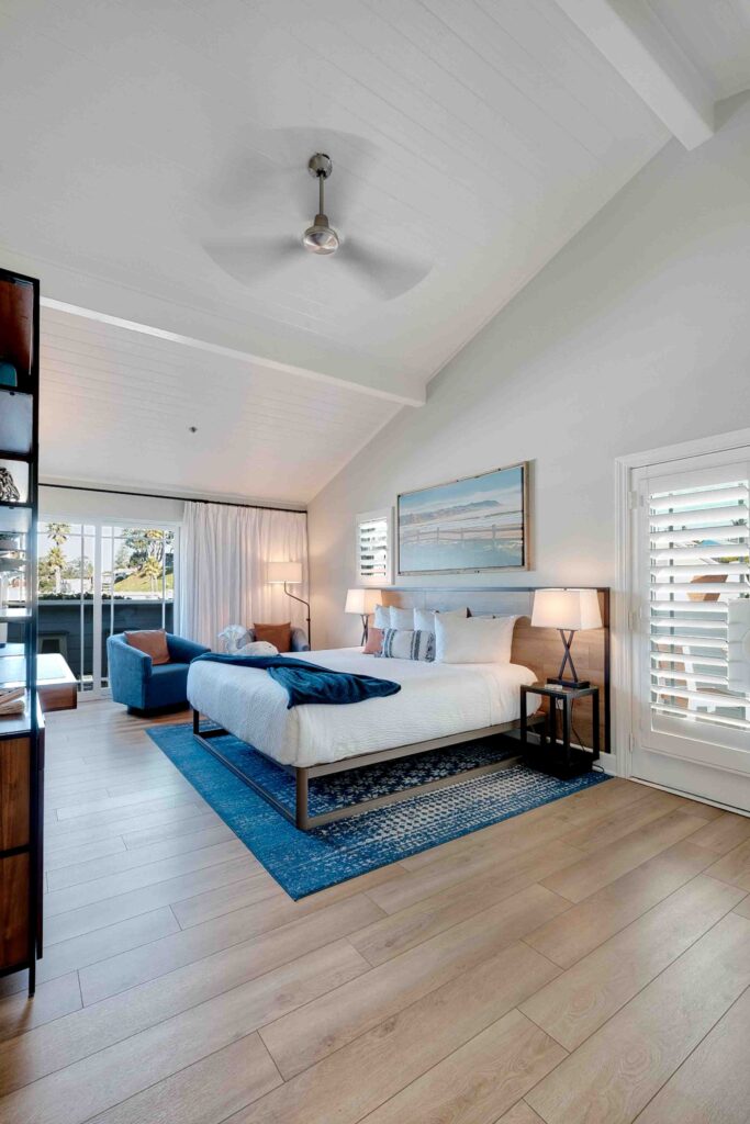 Modern bedroom with a high, white wood-paneled ceiling and a ceiling fan. The room features a large bed with blue accents, bedside tables with lamps, a blue armchair, a blue rug, and a wall-mounted painting. Large windows with white shutters offer a bright, airy feel.