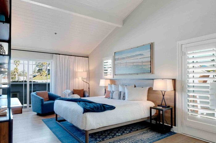 Modern bedroom with a high, white wood-paneled ceiling and a ceiling fan. The room features a large bed with blue accents, bedside tables with lamps, a blue armchair, a blue rug, and a wall-mounted painting. Large windows with white shutters offer a bright, airy feel.