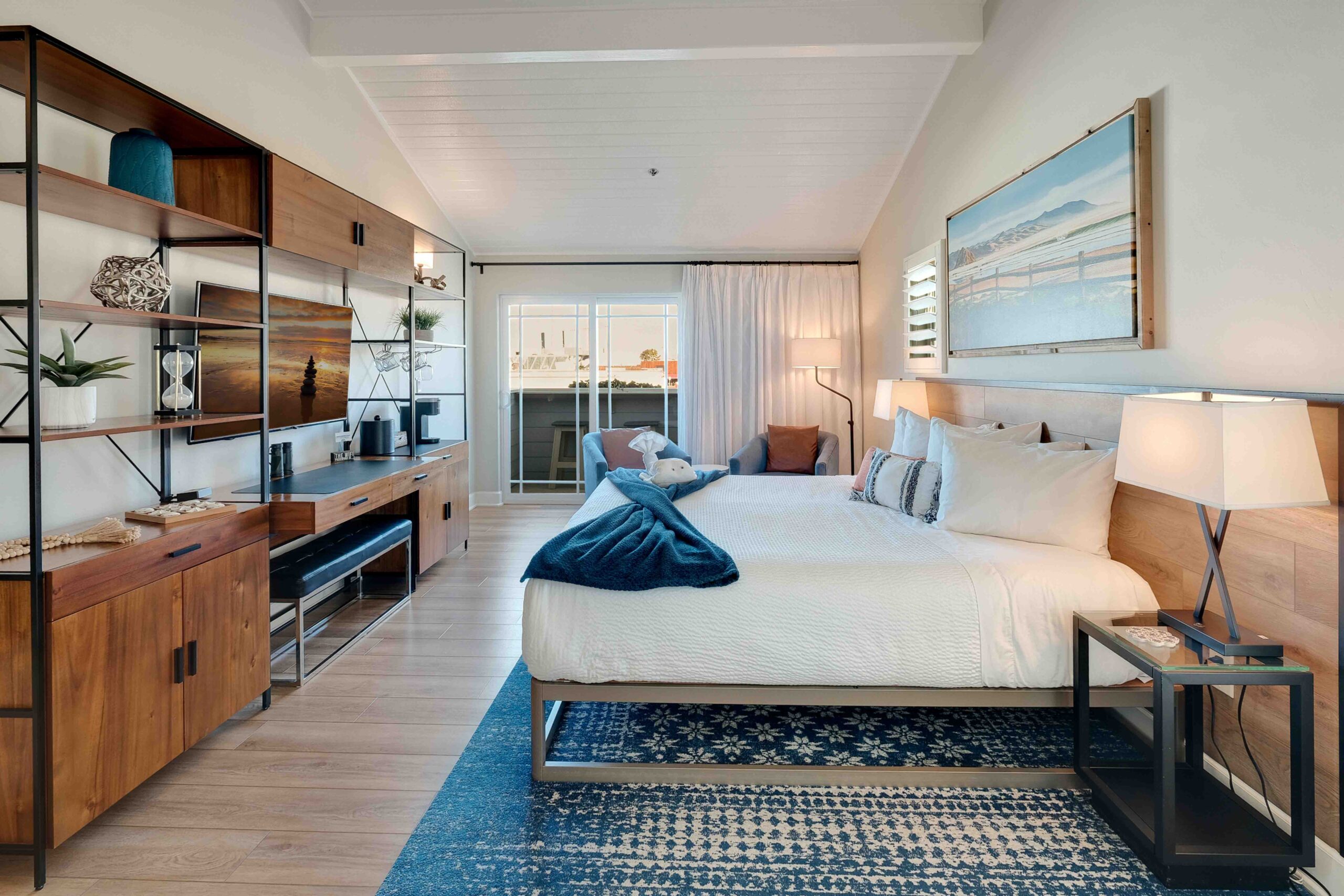 A modern, stylish bedroom features a large bed with white linens and blue accent pillows. Wooden shelving and storage units line the left wall. The room is brightened by natural light coming through a sliding glass door that leads to a balcony. A blue rug covers the wooden floor.