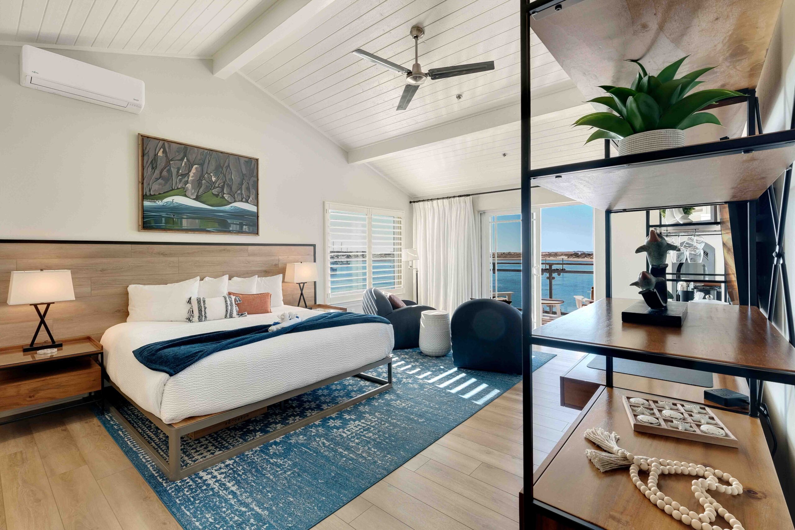 A modern bedroom with a large bed featuring white linens and a blue throw. There's wooden decor including a headboard and nightstands with lamps. The room has a wooden shelf with decorative items, chairs by the window, a ceiling fan, and a view of water through large windows.