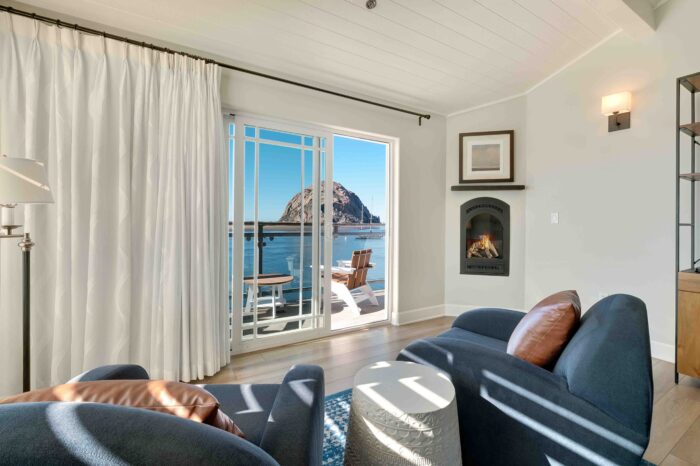 A cozy room with two dark blue armchairs facing a fireplace. A sliding glass door opens to a balcony with a table and chairs, offering a stunning view of a large rock formation in the ocean under a clear blue sky. White curtains and soft lighting create a serene atmosphere.