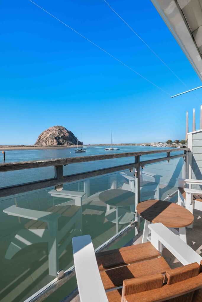 A wooden deck with chairs and a table offers a view of a bay with boats and a large rock formation in the background. The scene is under a clear blue sky, with water gently reflecting the landscape. Cable lines and part of a building's exterior are visible on the side.