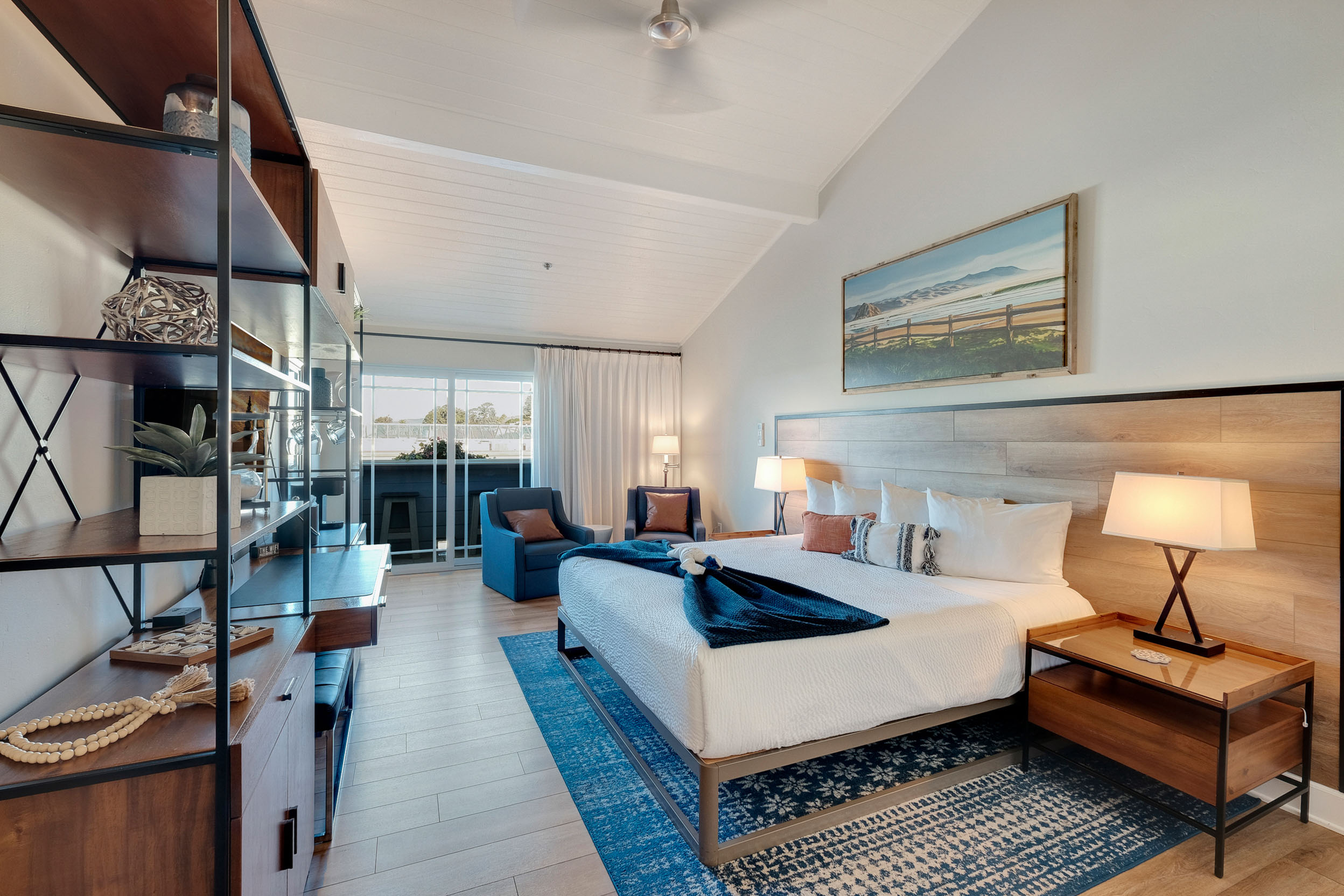 A cozy, well-lit bedroom with a large bed, two pillows, and a folded blanket. There are wooden nightstands with lamps on either side of the bed. A blue chair and table are by the window, with a view outside. A bookshelf with decor is on the left side of the room.