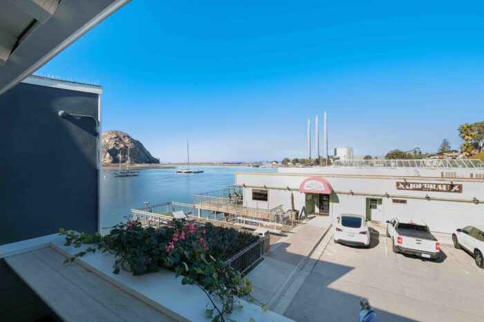 A scenic view from a balcony overlooking a calm bay with anchored sailboats, a building labeled "Hofbrau," and a large rock formation in the distance under a clear blue sky. Potted flowers are seen on the balcony railing. Two white vehicles are parked below.