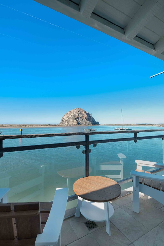 A serene view of Morro Rock across calm, blue waters from a balcony. The balcony has a glass railing and is furnished with white chairs and a round wooden table, all under a clear, blue sky. Sailboats are visible in the distance near the rock formation.