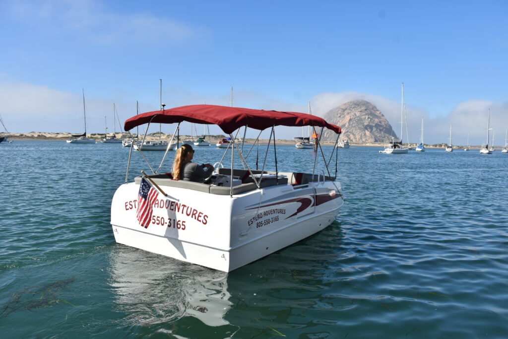 A small motorboat with a red canopy, labeled "ESTR ADVENTURES" and displaying a phone number, floats on calm water with several other boats anchored nearby. A large rock formation is visible in the background under a partly cloudy sky. An American flag is attached to the boat.