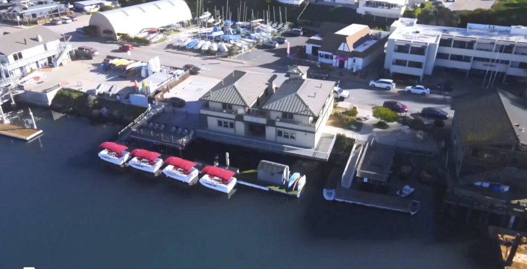 Aerial view of a marina with several red-roofed boats docked beside a large building. The area includes various structures, boats in storage, and parking spaces. The marina is surrounded by water and nearby buildings, with visible streets and vehicles.