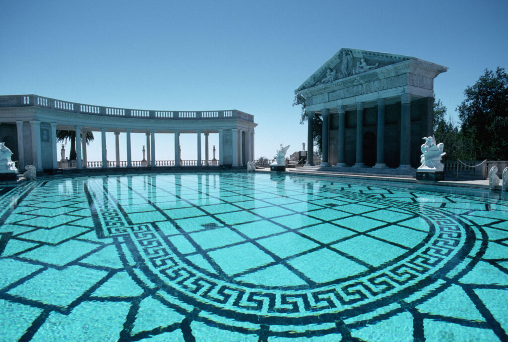 A luxurious outdoor pool featuring ornate tilework and clear, turquoise water. Surrounding the pool are neoclassical structures with columns and statues, including a temple-like building. The background showcases a clear blue sky and distant trees.