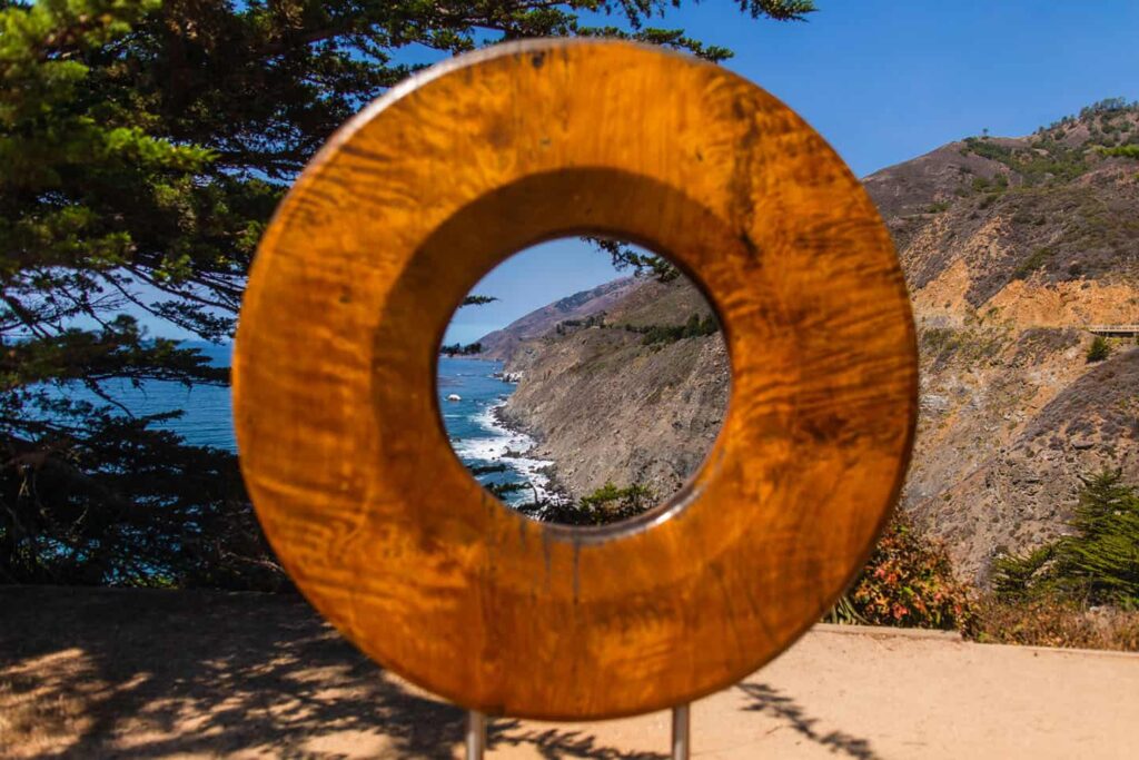 A large circular wooden sculpture with a hollow center stands outdoors, framing a picturesque coastal scene. Through the sculpture's opening, rugged cliffs, a winding road, and the ocean are visible under a clear blue sky. Trees flank the sculpture.