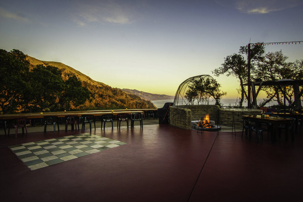 A serene outdoor patio with a chessboard pattern on the floor, overlooking a picturesque landscape of mountains and the ocean at sunset. There are tables and chairs around the area with a cozy fire pit surrounded by trees.