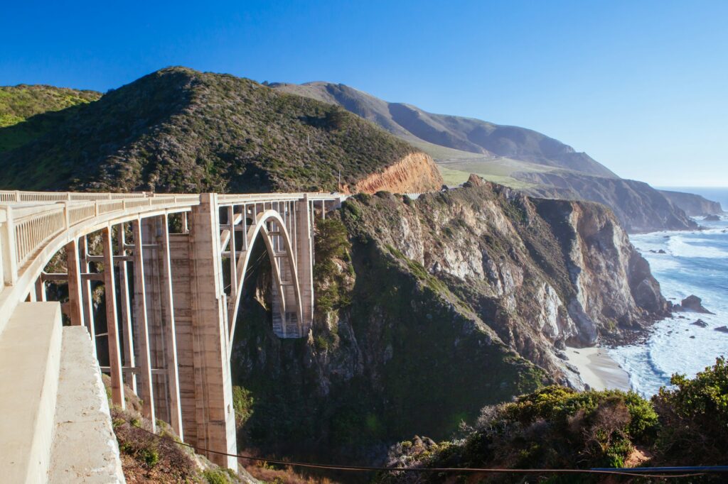 A scenic view of Bixby Creek Bridge along California's Big Sur coastline. The concrete arch bridge spans over rugged cliffs with lush hills in the background and waves crashing against the rocky shore below. The sky is clear with bright sunshine.