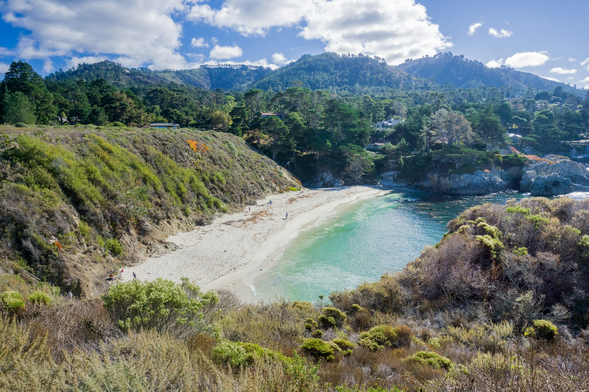 A scenic coastline features a secluded, sandy cove with clear, turquoise water surrounded by rugged cliffs and green foliage. In the background, a forested hillside rises under a sky filled with fluffy white clouds.