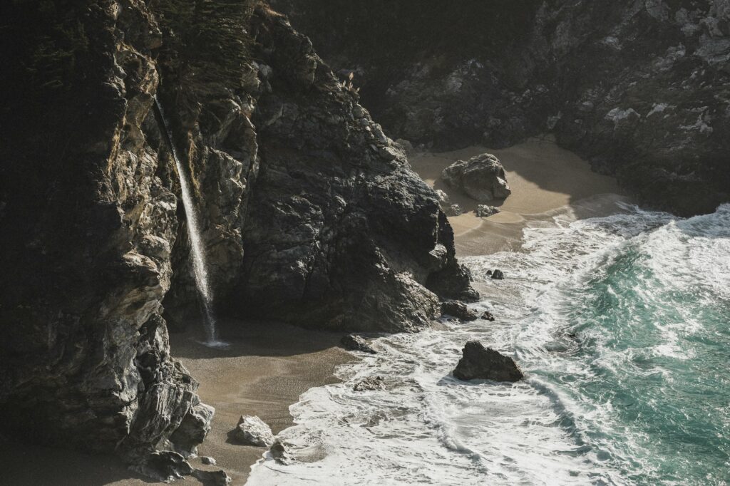 A secluded beach with a small waterfall cascading down rocky cliffs into the sandy shore. The turquoise waves gently lap against the shore, and the surrounding rocks and cliffs add to the rugged, natural beauty of the scene.